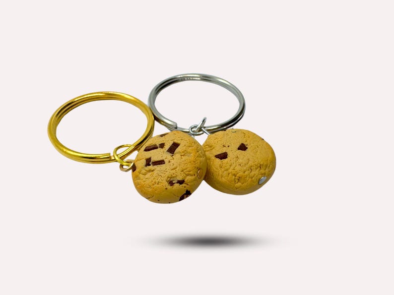 May include: Two chocolate chip cookie keychains, one on a gold key ring and the other on a silver key ring. The cookies are brown and have chocolate chips.