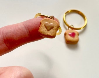 Puede incluir: Dos llaveros de oro con un dije de tostada en forma de corazón. Un dije tiene jalea rosa y el otro tiene un relleno marrón claro.