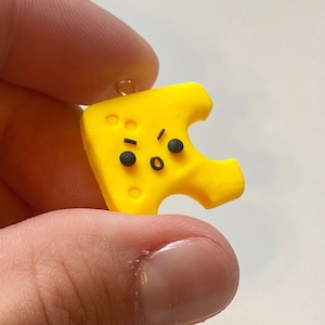 May include: A yellow cheese charm with a cartoon face and holes. The charm has a gold loop for hanging.