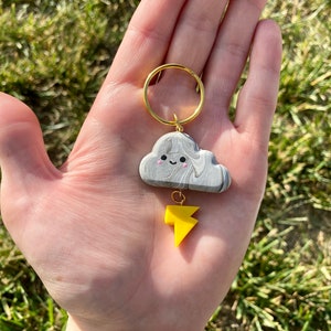 Puede incluir: Un charm de nube marmoleado gris y blanco con una cara sonriente y un charm de rayo amarillo unido a un llavero dorado.