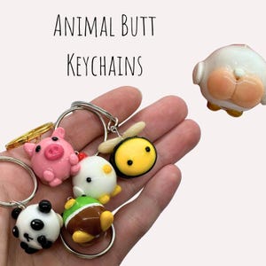 Può includere: Una collezione di portachiavi originali con disegni di sederi di animali. I portachiavi includono un maiale rosa, un panda, un'ape, un'anatra e un animale marrone e verde. Il testo "ANIMAL BUTT KEYCHAINS" è visualizzato sopra.