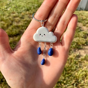 Puede incluir: Un llavero en forma de nube blanca con una cara sonriente y abalorios de gotas de lluvia azules.