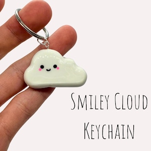 Peut inclure: Porte-clés en forme de nuage blanc avec un visage souriant et des joues roses. Le porte-clés est attaché à un anneau de clé argenté. "SMILEY CLOUD KEYCHAIN" est écrit en noir sous le nuage.