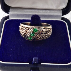 Könnte beinhalten: Ein goldfarbener Ring mit einem verzierten Filigran-Design. Der Ring hat drei smaragdgrüne Edelsteine, die in einem diagonalen Muster angeordnet sind. Der Ring wird in einer blauen Schmuckschatulle mit Samtinnenraum präsentiert.