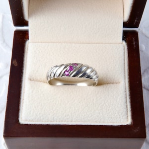 May include: A silver ring with a ribbed design and three small, bright pink gemstones set in the centre. The ring is displayed in a dark wooden jewellery box with a cream-coloured interior.