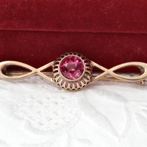 Op de afbeelding: Goudkleurige broche met een centrale, ronde, roze edelsteen. De edelsteen is gezet in een gedetailleerd, opengewerkt frame. De broche heeft een langwerpig, symmetrisch ontwerp met een speldsluiting.