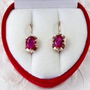 Op de afbeelding: Een paar goudkleurige oorbellen met ovale, magenta-kleurige edelstenen. De oorbellen worden gepresenteerd in een hartvormige rode doos met een wit interieur, tegen een witte, getextureerde achtergrond.