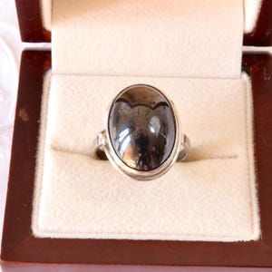 May include: A silver ring featuring a large, polished, oval-shaped dark grey hematite gemstone set in a smooth metal bezel. The ring is displayed inside a square, dark brown wooden jewellery box with a cream-coloured, soft velvet interior lining.