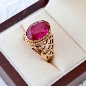 May include: A gold ring featuring a large, oval-cut, deep red gemstone. The ring has an openwork design on the band and is set in a gold bezel. The ring is displayed in a brown jewellery box with a cream interior.