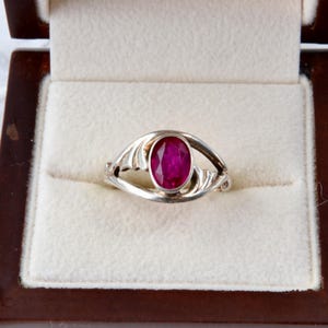 May include: A silver ring with an oval, deep red gemstone, presented in a wooden jewelry box with a cream-colored interior. The ring's design features flowing, stylized details around the gemstone.