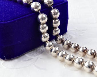 May include: A silver beaded necklace with large, round beads. The necklace is displayed next to a blue velvet jewellery box on a white, patterned surface. The beads are strung together, creating a simple yet elegant design.