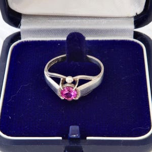 Könnte beinhalten: Ein silberfarbener Ring mit einem runden, facettierten, leuchtend pinkfarbenen Edelstein in einer Krappenfassung. Die Ringschiene hat ein geteiltes Design, das an dem Stein zusammenläuft. Der Ring liegt in einer dunkelblauen, samtgefütterten Schmuckschatulle.