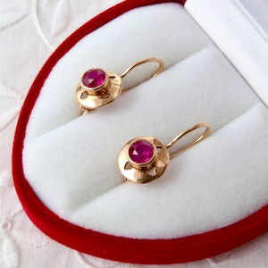 Puede incluir: Un par de pendientes de oro con una gema roja redonda en cada uno. Los pendientes se exhiben en un joyero de terciopelo rojo con interior blanco. Los pendientes tienen un diseño de gancho.