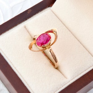 Pode incluir: Um anel de ouro com uma grande pedra preciosa vermelha em forma oval. O anel tem um design único com elementos curvos em forma de asa em ambos os lados da pedra preciosa. É exibido numa caixa de joias castanha com interior creme.
