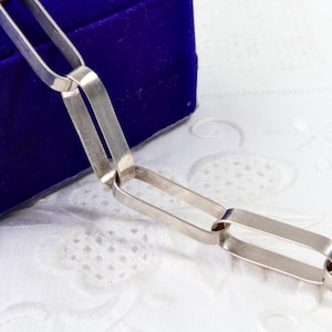 Può includere: Una collana a catena color argento caratterizzata da maglie rettangolari allungate con angoli arrotondati. La catena è composta da una serie di maglie metalliche piatte interconnesse che creano un motivo uniforme e continuo.