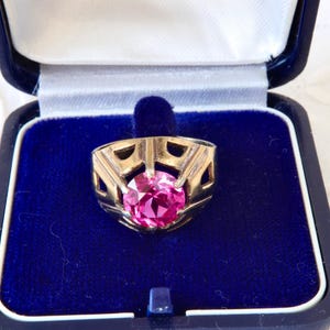 May include: A gold-toned ring featuring a large, round, faceted pink gemstone set within a geometric, open-work metal band. The ring is displayed inside a dark blue, hinged jewelry box with a plush, deep blue velvet interior lining.