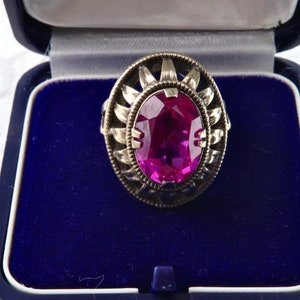 Può includere: Un anello vintage in argento con una grande pietra preziosa ovale color magenta. L'anello ha una montatura decorativa in argento con un design a raggiera. L'anello è presentato in un cofanetto blu.