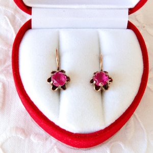 Peut inclure: Boucles d'oreilles dorées avec un motif floral, chacune ornée d'une pierre précieuse rose vif au centre. Les boucles d'oreilles sont présentées dans un écrin rouge en forme de cœur avec un intérieur blanc, sur un fond en tissu blanc.