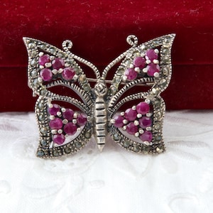 Può includere: Spilla a forma di farfalla argentata con pietre color rubino e dettagli in marcasite. La spilla presenta un design dettagliato con le pietre di rubino raggruppate nelle ali e nel corpo della farfalla.