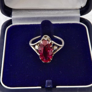 Puede incluir: Un anillo de plata con una gran piedra preciosa de color rojo intenso, de corte ovalado y engastada en garras. El aro tiene un diseño dividido con pequeñas esferas de plata a cada lado de la piedra central, presentado en un joyero azul.