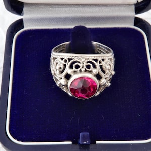 Op de afbeelding: Een zilveren ring met een sierlijk filigraanontwerp en een grote, ronde, rode edelsteen. De ring wordt gepresenteerd in een blauw fluwelen sieradendoosje met een witte bloemenachtergrond.