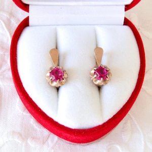 Puede incluir: Un par de pendientes dorados con un diseño floral, cada uno con una vibrante gema roja redonda. Los pendientes se exhiben en un joyero rojo en forma de corazón con interior blanco, sobre un fondo de tela blanca.