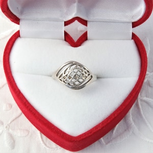 Puede incluir: Un anillo de plata con un diseño calado, presentado en una caja de joyería roja y blanca en forma de corazón. El anillo presenta un patrón central en forma de diamante dentro de un marco.