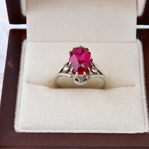May include: A silver ring with a large, oval-cut, vibrant red gemstone. The ring is set in a decorative setting and presented in a dark wooden jewelry box with a cream-colored interior. The ring is a piece of jewelry.