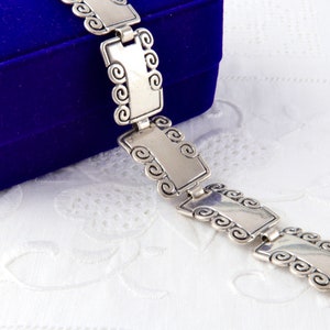 Puede incluir: Pulsera plateada con eslabones rectangulares y detalles decorativos en espiral. La pulsera se encuentra parcialmente sobre una caja de terciopelo azul, sobre un fondo blanco con diseño. Una joya clásica.