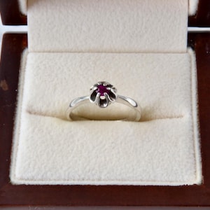 Può includere: Un anello d'argento con montatura a forma di fiore e una pietra preziosa rosso scuro. L'anello è esposto in un portagioie di legno con interno color crema.