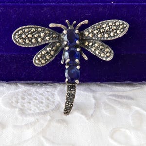 以下が含まれることがあります： 銀色のトンボのブローチ。羽にはマーカサイトのアクセント、胴体には青い宝石が並んでいます。ブローチは紫色のベルベットの背景に置かれています。