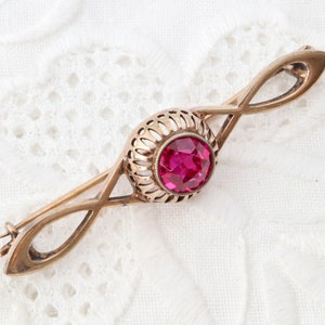 Può includere: Una spilla in metallo color oro con un design a barra allungata e intrecciata. Al centro, una gabbia circolare in stile filigrana racchiude una singola pietra preziosa rotonda, sfaccettata e di colore rosa intenso. Il metallo ha una finitura calda.