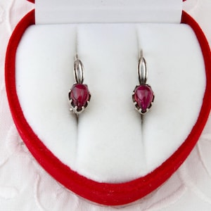 Op de afbeelding: Een paar zilveren oorbellen met druppelvormige, dieprode edelstenen. De oorbellen worden gepresenteerd in een hartvormige rode doos met een witte binnenkant, tegen een witte, getextureerde achtergrond.