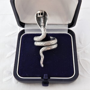 Peut inclure: Une bague argentée en forme de serpent cobra enroulé avec une finition de surface martelée et texturée. La tête du serpent est relevée avec des détails oculaires visibles, et le corps s'enroule pour former l'anneau, présentée dans un écrin en velours bleu.