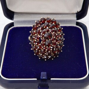 May include: An oval ring featuring a cluster of small, dark red gemstones set in a silver or gold band. The ring is presented in a blue velvet jewellery box. The gemstones are closely packed, creating a textured surface.