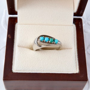 Puede incluir: Un anillo de plata con cinco piedras turquesas dispuestas en un diseño curvo. El anillo se exhibe en un joyero marrón oscuro con interior crema, resaltando el diseño elegante y el contraste de colores.