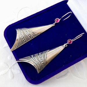 May include: A pair of silver dangle earrings featuring a triangular, curved shape with a textured surface of raised dots and lines. Each earring is accented with a small, round, deep pink gemstone set at the top near the hook closure.