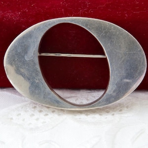 以下が含まれることがあります： 大きな中央の開口部がある銀色の楕円形のブローチ。ブローチはシンプルでエレガントなデザインで、裏側にピン留めが付いています。背景は赤いベルベットの表面と白いレース生地です。