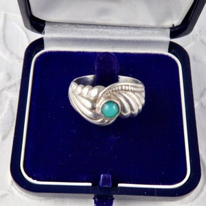 Peut inclure: Une bague en argent ornée d'une pierre polie et arrondie de couleur turquoise au centre. L'anneau présente un design texturé et nervuré qui s'incurve autour de la pierre, créant un aspect superposé. La bague est présentée dans un écrin bleu.