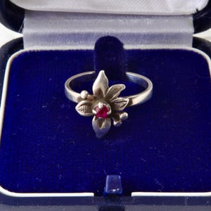 Può includere: Un anello in argento con un'impostazione a forma di fiore con petali testurizzati e una piccola pietra preziosa rotonda di un rosso vibrante al centro. L'anello è esposto all'interno di un portagioie blu scuro foderato in velluto con bordo bianco.