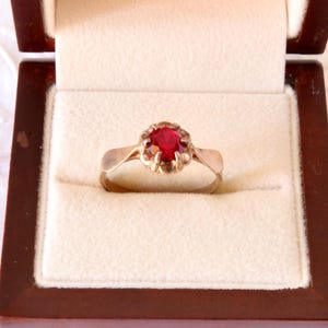 Könnte beinhalten: Ein goldener Ring mit einem runden, facettierten, tiefroten Edelstein in einer mehrzinkigen Fassung. Der Ring befindet sich in einer dunkelbraunen Holzschatulle mit cremefarbenem Samtfutter, die zur Aufbewahrung und Präsentation des Schmuckstücks dient.