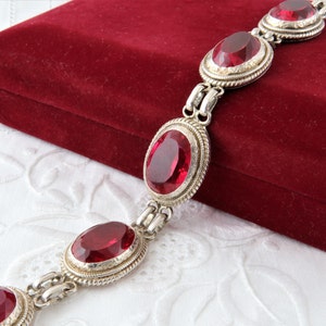 Puede incluir: Un brazalete de plata con piedras preciosas ovaladas rojas. El brazalete tiene un delicado diseño de cadena y se presenta sobre un fondo de terciopelo rojo.