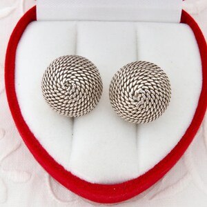 May include: Two silver-coloured stud earrings with a spiral rope design. The earrings are presented in a heart-shaped red and white jewellery box, set against a white, textured background.