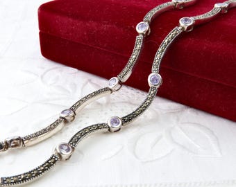 Può includere: Una collana d'argento dal design ondulato, con piccole pietre preziose rotonde, viola chiaro e pietre più piccole, grigio scuro. La collana è esposta su una superficie bianca a motivi con una scatola di velluto rosso sullo sfondo.