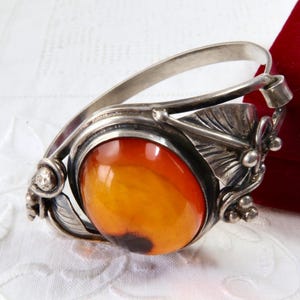 May include: A silver-toned bracelet with an oval, amber-colored stone. The bracelet features leaf and floral details around the stone. The background is a white, textured surface and a red box.