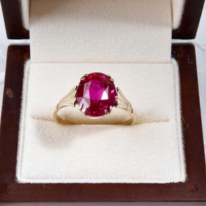May include: A gold ring featuring a large, oval-cut, deep red gemstone, displayed in a dark brown jewellery box with a cream interior. The ring has a classic setting.
