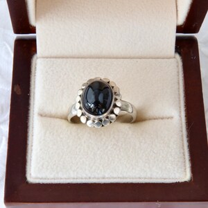 May include: A silver ring featuring a central oval black stone surrounded by a scalloped metal border. The ring is displayed inside a dark brown wooden box with a cream-coloured velvet interior lining, designed to hold jewellery securely in place.
