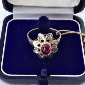 Può includere: Anello in argento con montatura a forma di fiore e una gemma rossa ovale al centro. L'anello è esposto in un cofanetto blu con interno in velluto. Un'etichetta è attaccata.
