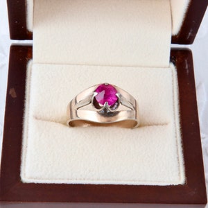 Puede incluir: Un anillo dorado con una gran gema roja brillante de forma ovalada en el centro. El anillo se muestra dentro de un joyero de madera con un interior color crema.