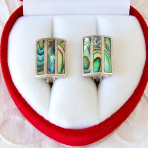 Può includere: Un paio di gemelli rettangolari in argento con intarsi di conchiglia di abalone iridescente, presentati in una scatola rossa a forma di cuore. La conchiglia di abalone presenta sfumature di verde, blu e viola.